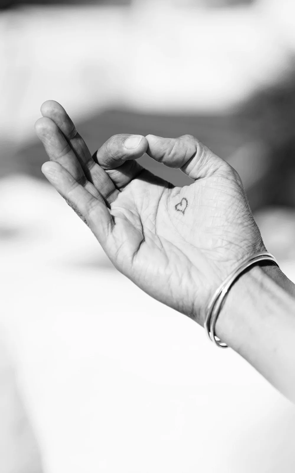 Hand in Mudra-Haltung mit kleinem Herz-Tattoo und silbernen Armreifen, schwarz-weiß